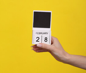 Female hand holding wooden calendar with date February 28 on yellow background