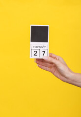 Female hand holding wooden calendar with date February 27 on yellow background