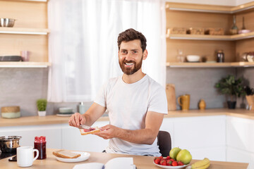 Fototapeta na wymiar Cheerful middle aged caucasian male with beard in white t-shirt makes sandwich with fruits for breakfast