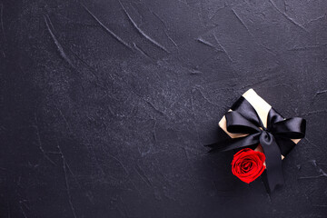  Romantic postcard. Wrapped box with present  and bright red rose flower on black textured background. Place for text. - 550666801