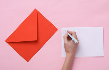 Female hand writes a letter on a pink background with a red envelope