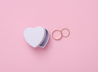 White heart shaped box with gold wedding rings on pink background. Top view