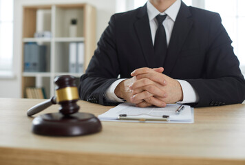 Cropped shot of professional lawyer or attorney in suit sitting at wooden table with judge's gavel and paper documents. Legal services and law consultation concept background