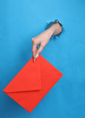 Female hand holds red envelope through torn hole in blue paper