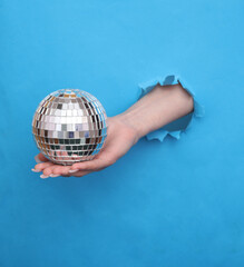 Female hand holds mirror disco ball through torn hole of blue paper