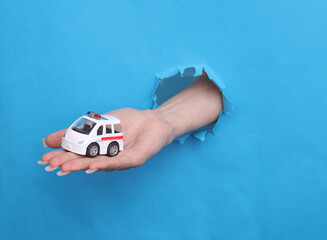 Female hand holds toy ambulance car through torn hole of blue paper