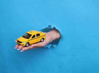 Female hand holds toy taxi car through torn hole of blue paper