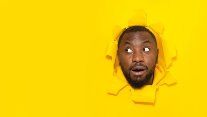 Omg. Shocked black man looking aside at free space through hole in torn yellow paper, posing with opened mouth, panorama