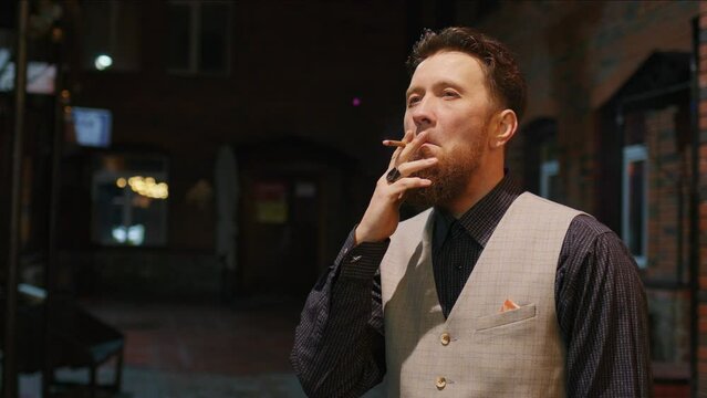 Red-haired middle-aged man having cigarette in yard of brick house. Bearded guy takes puff and lets out smoke, standing on dark street. Smoker doing smokebreak, thinking about something outdoor