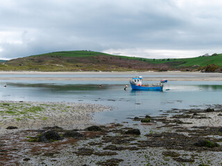 Obraz premium A small old fishing boat is anchored, shallow water at low tide on a cloudy day. Hills under a cloudy sky.