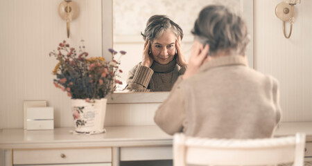 Beautiful old woman sits near the mirror in her cozy light room.