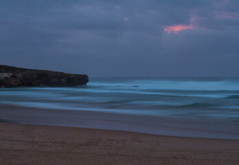 Mysterious moody long exposure view of sand beach Praia da Amoreira beach with with blurred ocean waves and pink and blue clouds. Blue hour after sunset at Rota Vicentina coast, Almograve, Portugal.