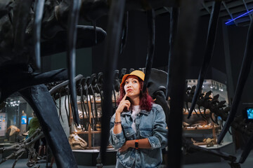 Excited tourist girl at the excursion looks thoughtfully at the fossilized remains of a dinosaur skeleton in the Museum of Natural History.