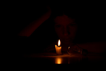A girl in the dark sits with candles and a light bulb at a table in the dark at home, people...