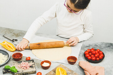 Caucasian girl in a white turtleneck with glasses rolls out pizza dough with a rolling pin .