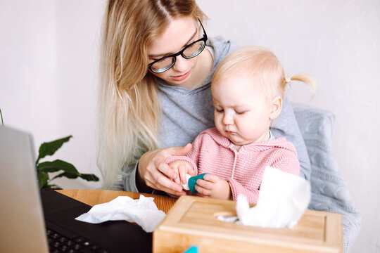 Portrait of young mother sitting at table near laptop, holding little baby, looking at bottle with medicine at home.