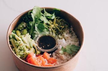 Poke bowl with sushi rice, raw salmon, avocado, seaweed salad, rocket, edamame beans and spring onion with a soy sesame dressing in take away paper bowl