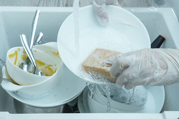 Fototapeta na wymiar Human hands in protective domestic gloves wash white plate with sponge under tap water. Dish washing, tableware after meal, food leftovers. Pile of kitchen utensils in sink closeup. Home cleaning.