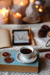 Cup of tea, books, tablet, bowl of cookies, various spices, pine cones and lit candles. Hygge at home. Selective focus.