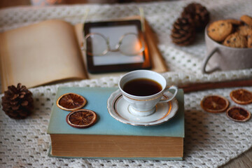 Cup of tea, books, tablet, bowl of cookies, various spices, pine cones and lit candles. Hygge at home. Selective focus.