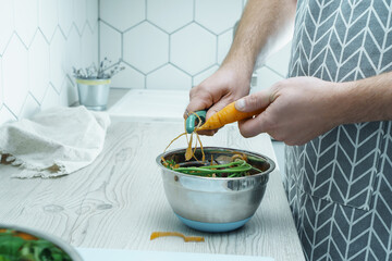 Male hands peeling fresh carrot with peeler over bowl of vegetable peelings. Cook on kitchen table, prepare ingredients, food scrap. Healthy nutrition, vegetables. Separate waste collection.