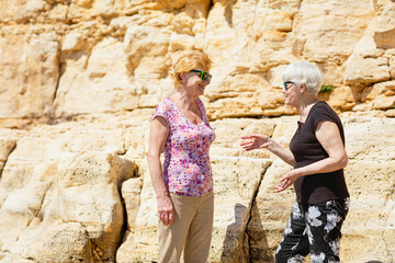 Two senior women are happy to meet each other, standing near rock, talking and laughing together. Elderly women are happy to communicate