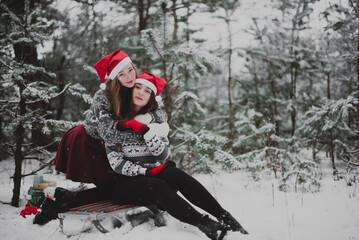Fototapeta na wymiar Two young teenage hipster girl friends together.Close up fashion portrait of two sisters hugs and having fun winter time,wearing red santa hats and sweater,best friends couple outdoors, snowy weather
