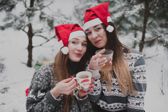 Close up fashion portrait of two sisters hugs and having fun, drinking tea winter time,wearing red santa hats and sweater,best friends couple outdoors, snowy weather