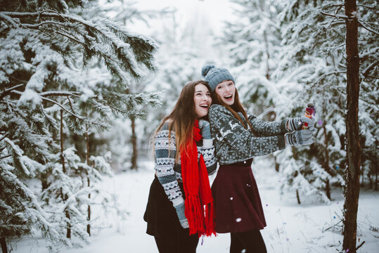 Close up fashion portrait of two sisters hugs and having fun, holding petard in winter time forest, wearing sweaters and scarfs,best friends couple outdoors, snowy weather