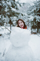 young teenage hipster girl having fun, make snowman in winter time forest, wearing sweaters and scarfs, outdoors, snowy weather