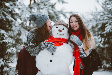Close up fashion portrait of two sisters hugs and having fun, make snowman in winter time forest,...