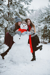 Close up fashion portrait of two sisters hugs and having fun, make snowman in winter time forest, wearing sweaters and scarfs,best friends couple outdoors, snowy weather