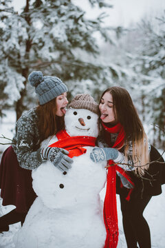 Close up fashion portrait of two sisters hugs and having fun, make snowman in winter time forest, wearing sweaters and scarfs,best friends couple outdoors, snowy weather