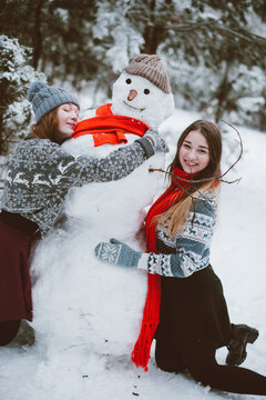 Close up fashion portrait of two sisters hugs and having fun, make snowman in winter time forest, wearing sweaters and scarfs,best friends couple outdoors, snowy weather