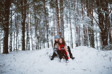 Fototapeta na wymiar Close up fashion portrait of two sisters hugs and having fun, ride on sled in winter time forest, wearing sweaters and scarfs,best friends couple outdoors, snowy weather