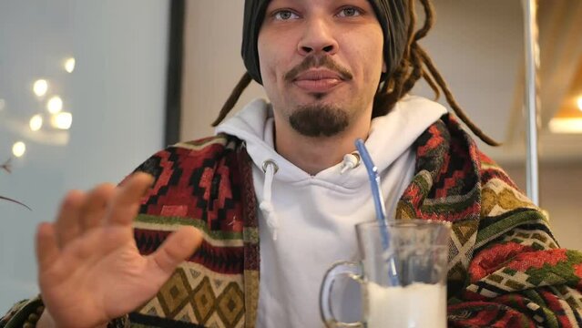 Attractive young mixed race man with dreadlocks and beard wrapped in a blanket with subculture patterns sitting at a cafe table eating ice cream with a spoon and talking. Handsome stylish rastafarian