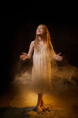 A girl in a white shirt and with long hair, looking like a ghost. A young model at a photo shoot with smoke, fog, flour, dust. A little fairy girl on a dark black background