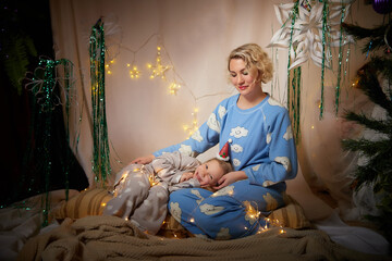 Cute mother and daughter in pajamas having fun in the room with Christmas garlands and white background. The tradition of decorating the house for the holidays. Happy childhood and motherhood