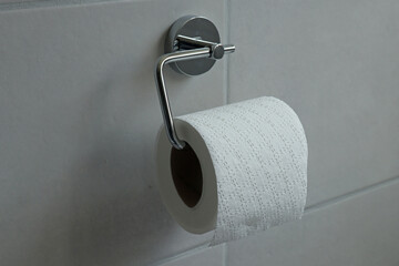 Close up of a chrome toilet roll holder with a white toilet roll in a grey tiled bathroom