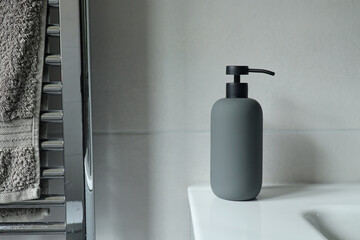 Grey soap dispenser on the sink in a grey tiled bathroom. A towel hangs from a chrome radiator