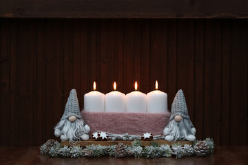 Four white burning candles with Christmas santa clauses in front of a wooden wall with space for text.