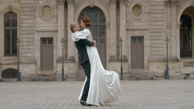 Happy just married groom carry on his hands the bridge in Paris, France. Action. Man in suit and woman in log wedding dress on the old architecture building background.