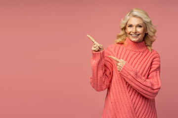 Smiling mature woman wearing stylish winter sweater pointing fingers on copy space, isolated on pink background. Shopping, sales concept 