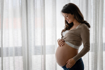 Portrait of a beautiful young pregnant woman standing by a big window and holding her belly. Always love your body. - obrazy, fototapety, plakaty