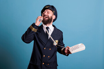 Middle aged airliner captain talking on landline phone, holding telephone, having conversation. Smiling pilot in professional flight uniform answering call in airport, studio medium shot