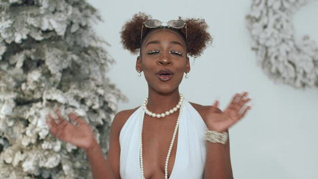 Attractive woman in holiday attire with open arms blows kiss in front of Christmas trees