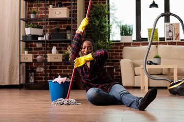 Stressed woman cleaning work sitting on the floor with no strength to continue with clean routine. Tired housewife holding mop looks away with negative expression of exhaustion.