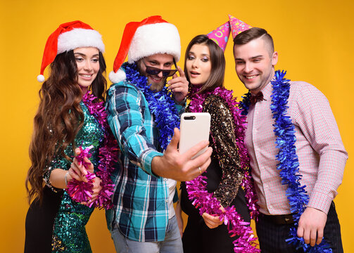 group or company of friends in santa hat, party hats, boas celebrating Christmas, having fun, taking selfies and going live on a yellow background.