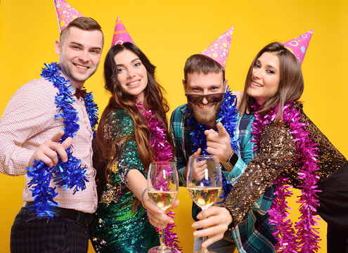 group or company of friends in party hats and colorful boas having fun and champagne glasses on a yellow background