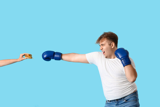 Young overweight man in boxing gloves fighting burger on blue background